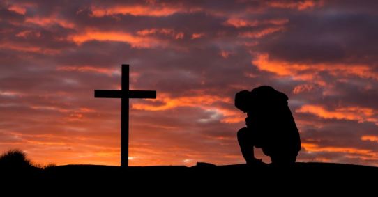 Man praying by a cross as the setting sun light up the clouds.